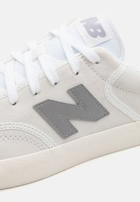 Baskets blanches en daim et mesh, avec un logo "N" en cuir gris, des lacets blancs et une semelle en caoutchouc texturée.