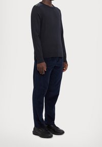 Navy long-sleeve sweater with a crew neck and colorful stripes on the left shoulder, paired with navy trousers and black sneakers.