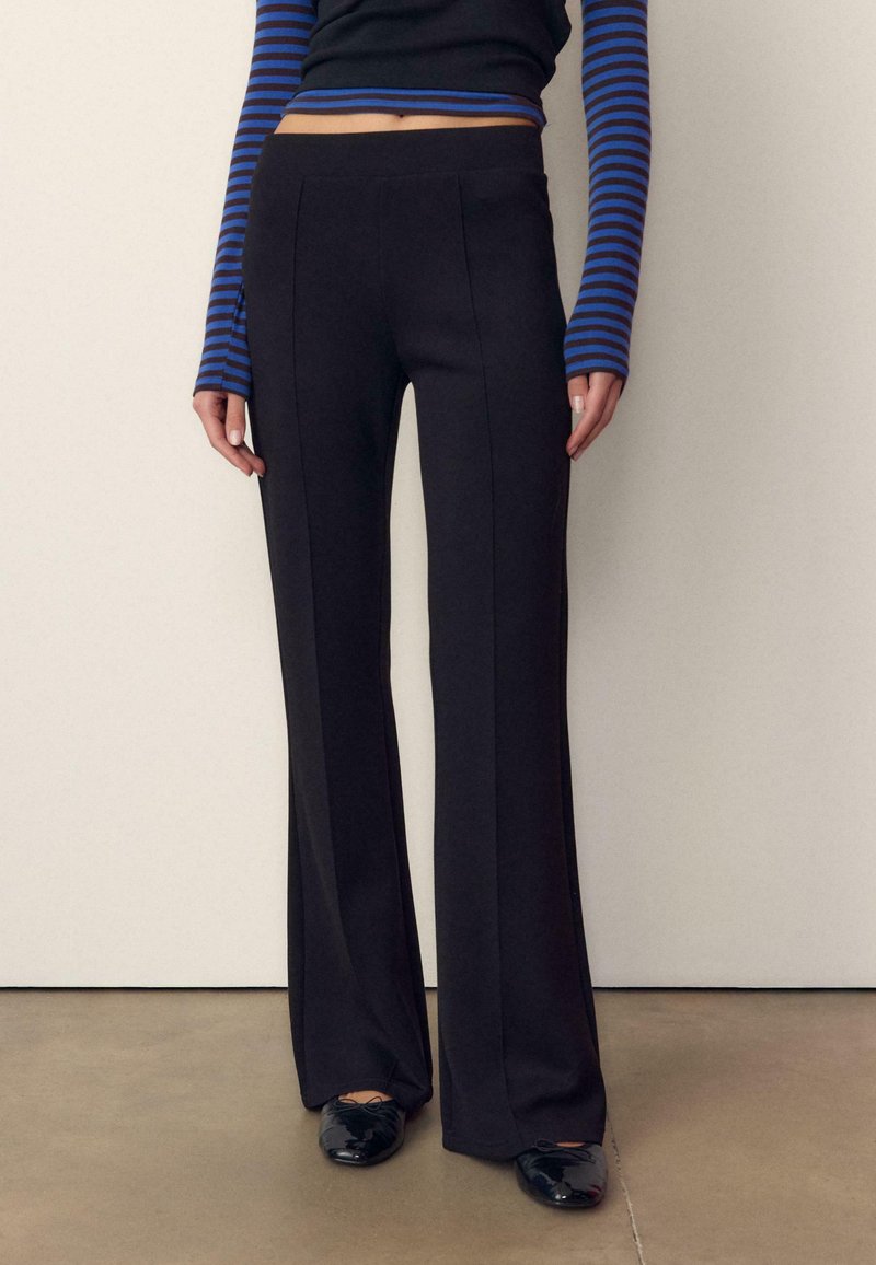 Persona che indossa pantaloni neri a gamba larga e vita alta, un top corto a maniche lunghe a righe blu e nere, e ballerine nere lucide.