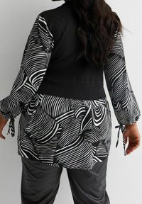 Frau, die eine langärmlige Bluse mit schwarz-weißer Musterung, einen schwarzen Strickwesten und schwarze Kunstlederhosen trägt und von der Kamera abgewandt ist.