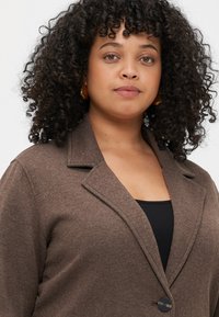 Blazer castanho em tecido trançado com lapelas recortadas, fecho de botão simples e tecido texturizado. Top preto visível por baixo. Brincos de argola dourada.