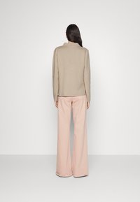 Vrouw met de rug naar de camera, gekleed in een beige langemouwen top en lichtroze, losse broek, tegen een plain witte achtergrond.