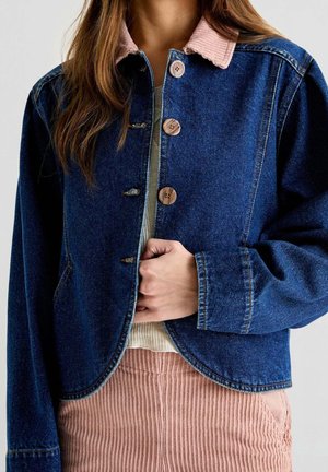 Kobieta nosząca niebieską kurtkę jeansową z drewnianymi guzikami i różowym kołnierzem ze sztruksu, w połączeniu z różowymi prążkowymi spodniami.