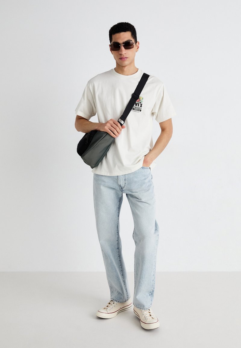 Camiseta de color beige claro con un pequeño gráfico, jeans rectos azules, bolso de cintura negro y zapatillas blancas. Gafas de sol de montura delgada completan el look.