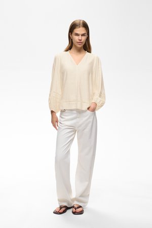 Jeune femme portant un chemisier ample crème, un pantalon blanc à jambes larges et des tongs foncées, debout avec une main dans la poche sur fond blanc.