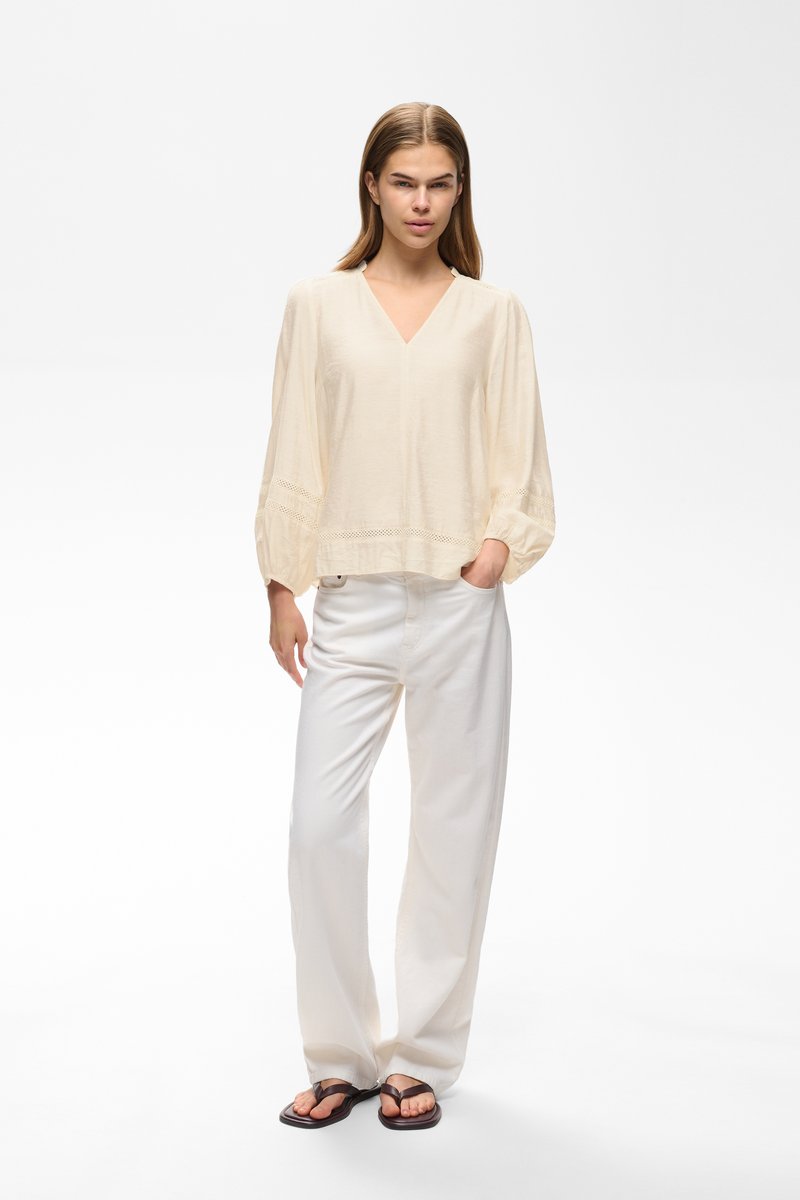 Jeune femme portant un chemisier ample crème, un pantalon blanc à jambes larges et des tongs foncées, debout avec une main dans la poche sur fond blanc.