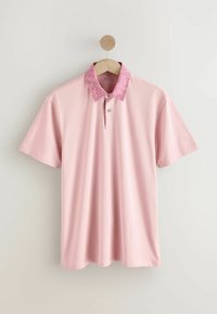 Polo-paita vaaleanpunaisena, jossa on kuvioitu vaaleanpunainen kaulus ja nappilista edessä. Valmistettu sileästä kankaasta, jossa on lyhyet hihat.