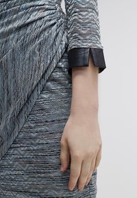 Tričko s dlouhými rukávy z texturované šedoblé fabrici s černým koženým manžetou, které se vyznačuje volným designem s jemnými vlnitými vzory.