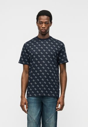 Ung mann iført en marineblå kortermet skjorte med mønster og blå jeans, stående mot en enkel hvit bakgrunn.