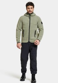 Grön fleecejacka med dragkedja fram, justerbar huva och två sidofickor. Svarta detaljer vid dragkedjor och ärmslut, bärs över mörka byxor.