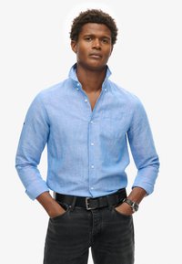 Camicia di lino azzurro chiaro con colletto button-down, maniche lunghe, taschino sul petto e una trama sottile, abbinata a jeans scuri e una cintura nera.