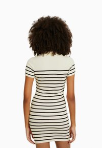 Femme aux cheveux bouclés portant une robe crème à manches courtes avec des rayures horizontales noires, vue de dos sur un fond blanc.