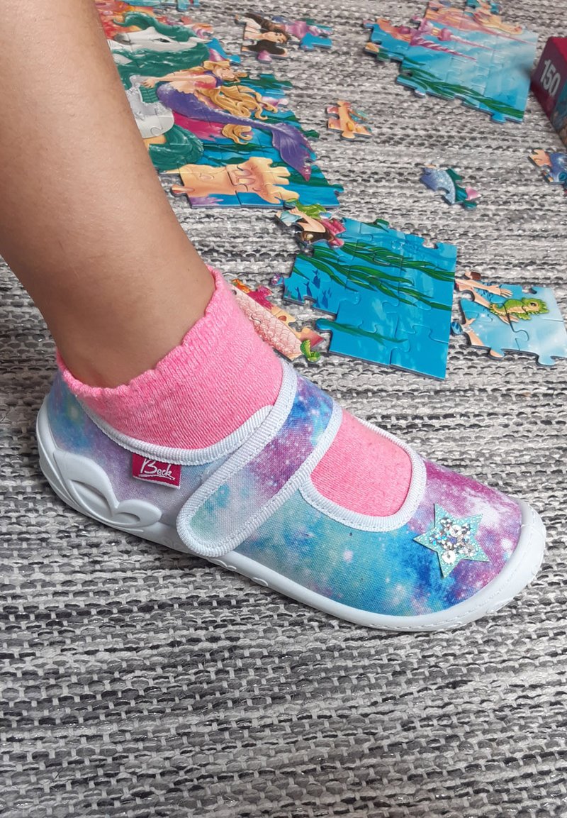 Kleurrijke canvas schoen met een sterrenhemelpatroon, witte zool en een blauwe steraccent. Roze sok zichtbaar boven de enkel, puzzelstukken op de achtergrond.