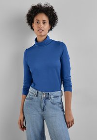 Blaues langärmeliges Rollkragenshirt aus glatter Stoffqualität, kombiniert mit hellblauen hochgeschnittenen Jeans mit doppeltem Knopfverschluss.