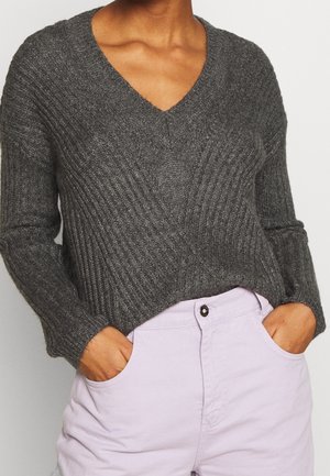 Personne portant un pull en côtes gris foncé à col en V et un pantalon lavande clair, les mains dans les poches, sur un fond uni.