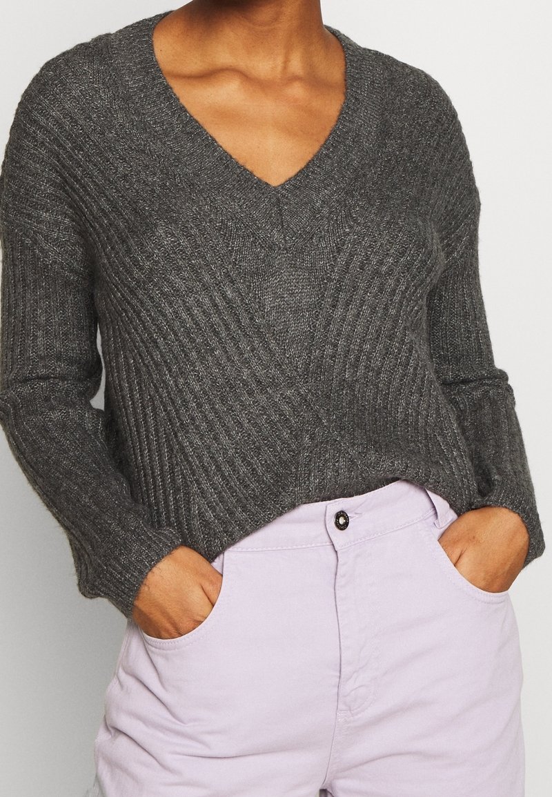 Personne portant un pull en côtes gris foncé à col en V et un pantalon lavande clair, les mains dans les poches, sur un fond uni.