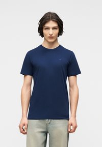 Νεαρός άνδρας με σκούρα μαλλιά μέχρι τους ώμους, φοράει απλό μπλε σκουρόχρωμο κοντομάνικο μπλουζάκι και ανοιχτόχρωμα τζιν, κοιτάζοντας προς τα εμπρός.