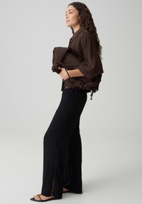 Woman with long curly hair wearing a brown shirt and black wide-leg pants, holding a large folded brown bag against a plain background.
