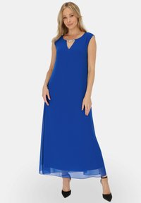 Kobieta ubrana w bezrękawnikową, królewską niebieską suknię maxi z ozdobnym dekoltem z kamieniami, w połączeniu z czarnymi szpilkami z zaostrzonymi noskami, stojąca na białym tle.