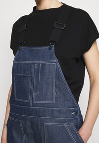 Džínové lacláče v tmavě modré barvě s kontrastním bílým prošíváním, nastavitelnými černými popruhy a několika kapsami pro úložný prostor. Klasický střih.