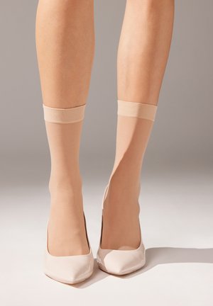 Beige knähöga strumpor med en slät yta och elastisk kant, bärs med blanka, spetsiga, ljusrosa högklackade skor. Den unika designen framhäver benen.