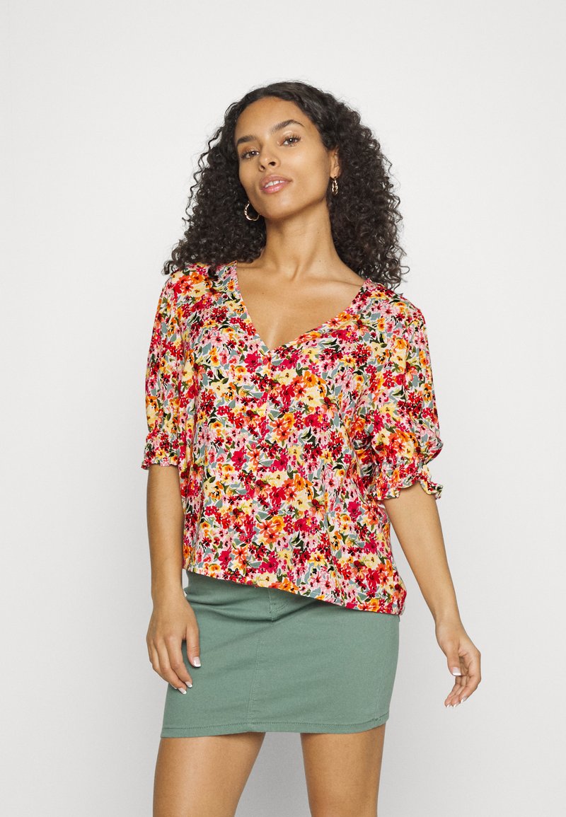 Blusa floral com decote em V, mangas curtas e punhos franzidos. Apresenta padrões em vermelho, amarelo e verde sobre um fundo claro. Combinada com uma saia verde.