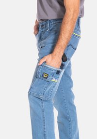 Pantalons de travail en denim bleu clair avec une poche latérale, dotés d'un accent jaune et d'un patch logo. Coupe droite avec des détails de couture visibles.