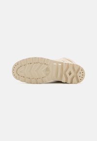 Semelle de chaussure beige avec un motif de grip texturé, featuring des lignes circulaires et ondulées. Fabriquée en caoutchouc durable ; plate avec un bout arrondi.