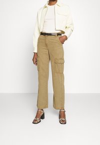 Veste jaune raccourcie portée sur un haut blanc, associée à un pantalon cargo marron avec poches latérales et ceinture noire. Les chaussures ont un motif serpent.