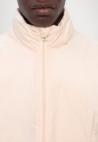 Lichtbeige ritsjas met hoge kraag, zachte textuur en minimalistisch ontwerp. Heeft een rits met een kleine trekker.