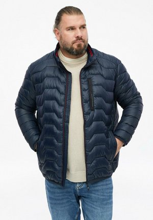 Mann mit Bart, der eine marineblaue gesteppte Jacke, einen beigen Pullover und blaue Jeans trägt, steht mit den Händen in den Taschen und schaut zur Seite.