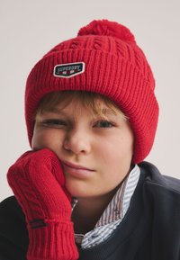 Rød strikket hue med pompon og mærkat, kombineret med matchende røde strikkede vanter. Begge er varme og har tekstur. Blå- og hvide stribede krave er synlig.