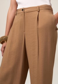 Tan wijde broek met een gestructureerde afwerking, voorplooien en een zwarte knoopsluiting. Voorzien van zijzakken en een relaxte pasvorm.