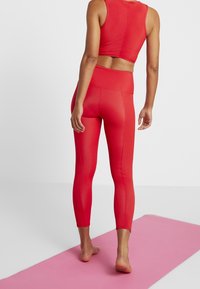 Czerwony sportowy top z krótkim rękawem i legginsy, oba z teksturowanej tkaniny. Legginsy mają wysoki stan i długość do kostek, ustawione na różowej macie.