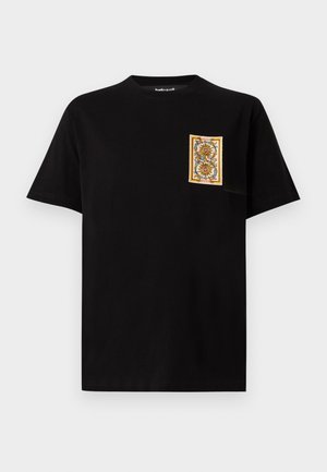 Camiseta negra de manga corta con cuello redondo, con un pequeño parche rectangular floral en amarillo, naranja y verde en el pecho izquierdo.