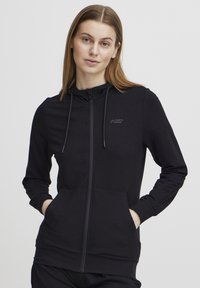 Black zip-up hoodie in soft fabric featuring a drawstring hood, front pockets, and subtle branding on the left chest area.