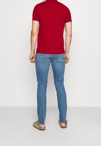 Chemise polo rouge à manches courtes assortie à un jean slim bleu clair et des mocassins beiges, montrant une tenue décontractée vue de dos.