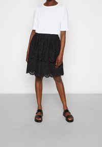 T-shirt blanc à manches courtes associé à une jupe en dentelle noire avec des bords en festons superposés et un design texturé, portée avec des sandales noires.