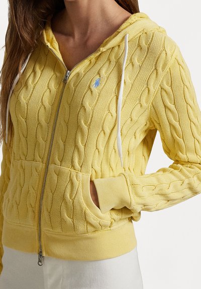 Geel kabelgebreide hoodie met een rits aan de voorkant, witte koordjes en een klein blauw logo. Heeft ribgebreide manchetten en een buidelzak.