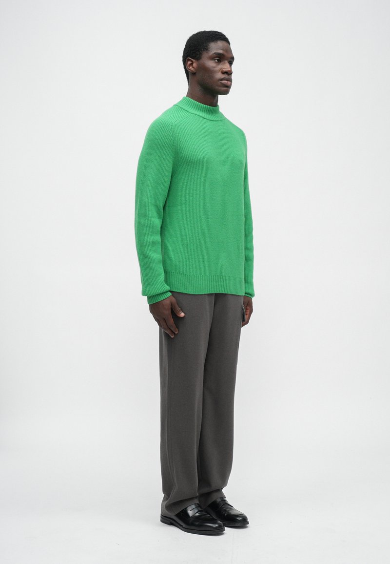 Pull en maille côtelée vert avec un col haut, porté avec un pantalon gris ample et des chaussures noires. Fond uni, posture debout.