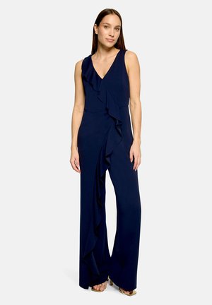 Žena nosi tmavomodrý overal bez rukávov s hlbokým výstrihom do V a volánikmi vpredu, v kombinácii s sandálmi na opätku s otvorenou špičkou.
