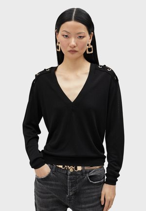 Black knit sweater with a V-neck and long sleeves, featuring gold-tone hardware accents on shoulders and ribbed cuffs. Paired with denim.