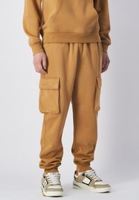 Beige Baumwoll-Sweatpants mit Bündchen an den Knöcheln, ausgestattet mit zwei großen Cargo-Taschen. Kombiniert mit grau-weißen Sneakers mit orangefarbenen Akzenten.