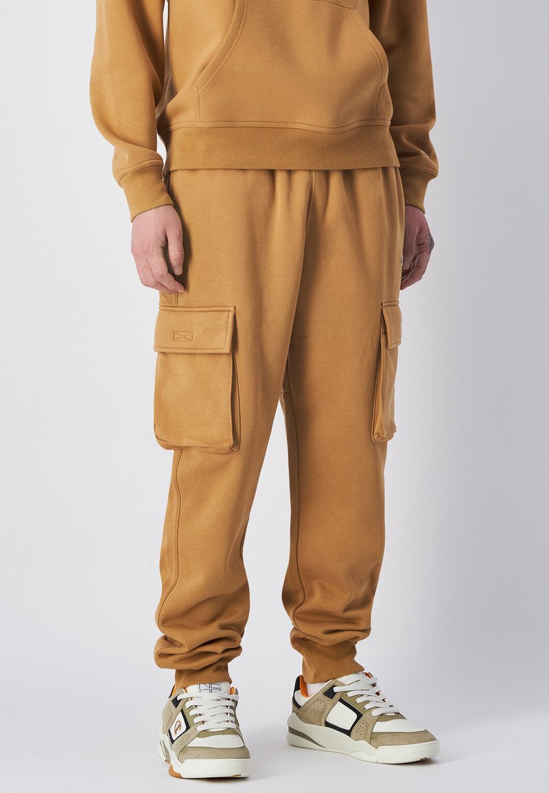 Beige Baumwoll-Sweatpants mit Bündchen an den Knöcheln, ausgestattet mit zwei großen Cargo-Taschen. Kombiniert mit grau-weißen Sneakers mit orangefarbenen Akzenten.