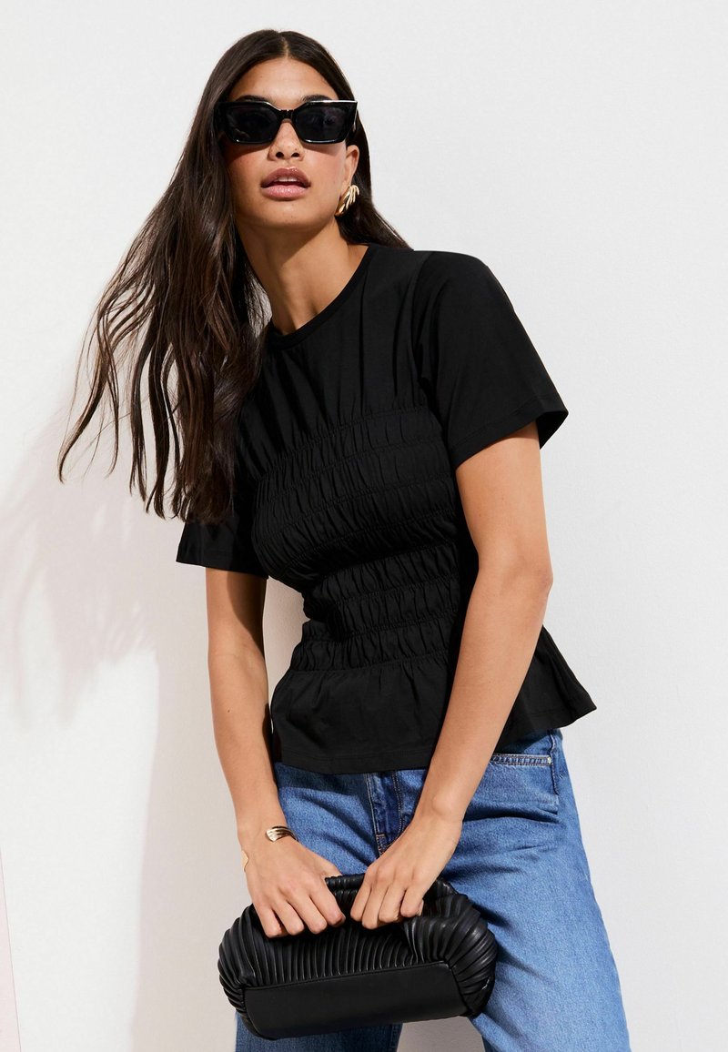 Črna zbrana majica z silhueto pepluma, kombinirana z modrimi kavbojkami z visokim pasom in teksturirano črno torbico. Poudarjena z naočniki v stilu mačjih oči.