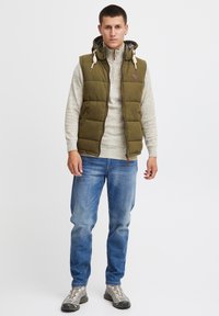 Gilet rembourré vert avec capuche sur un pull gris clair, associé à un jean bleu et des chaussures grises, avec des accents de zip et de cordon de serrage.