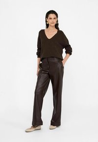 Brown knitted sweater with a V-neck, paired with high-waisted dark brown leather trousers. Model wears light-colored flats. Simple backdrop.