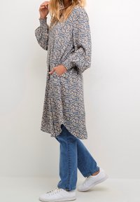 Vestido de estampado floral en beige y azul con mangas largas, escote en V y bolsillos, llevado sobre unos jeans de azul claro y zapatillas blancas.
