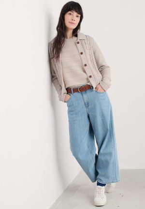 Beige kabelgebreide cardigan met houten knopen over een gestructureerde beige trui, gecombineerd met lichtblauwe wijde jeans en witte sneakers.