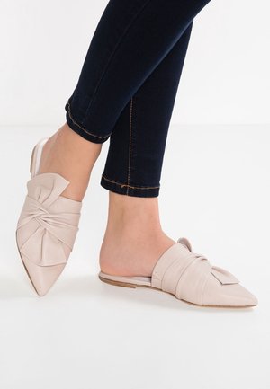 Pies con suaves planos de punta beige claro que se deslizan, con detalle de lazo, combinados con jeans oscuros ajustados sobre un fondo blanco.